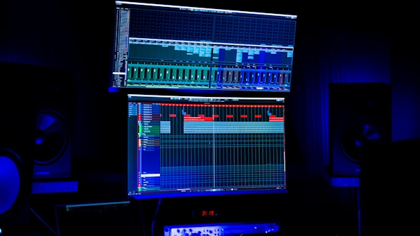 A dimly lit room features two large computer screens displaying complex audio editing software. Multiple speakers are visible in the background, with a digital clock and other electronic equipment situated below the screens.