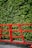 A red metal railing with geometric patterns stands in front of a lush green hedge. The vivid colors contrast sharply, enhancing the natural greenery against the man-made structure.