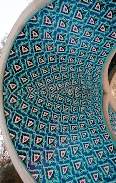 An intricately designed dome with a repeating geometric pattern featuring blue and turquoise hues. The design incorporates symmetrical shapes enclosed within a circular structure, hinting at Middle Eastern or Islamic architectural influences.