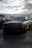 A black Mercedes-Benz race car is parked in a garage area beside other vehicles. The car features prominent AMG branding and a racing body kit. The sky is overcast with clouds, casting a moody light over the scene.