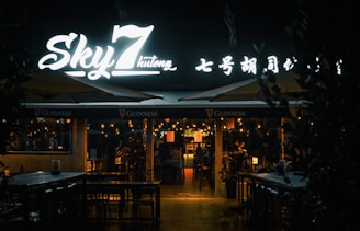 An outdoor seating area of a restaurant or bar is depicted at night. The establishment has a prominent illuminated sign that reads 'Sky 7 Hutong' along with some characters in another language. The area is lit with string lights, giving it a warm and inviting atmosphere. Tables and chairs are arranged neatly, and some umbrellas bearing the Guinness logo can be seen.