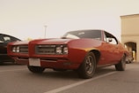 A classic red muscle car with a sleek design is parked in an outdoor lot. Its chrome details and vintage aesthetic stand out against the subtle background of a modern building.