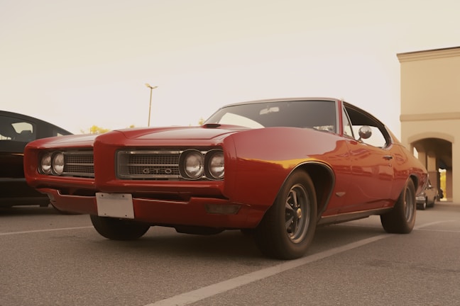 A classic red muscle car with a sleek design is parked in an outdoor lot. Its chrome details and vintage aesthetic stand out against the subtle background of a modern building.