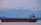 A large cargo ship is sailing across a calm body of water with a backdrop of snow-capped mountains and a hazy sky. The vessel is dark with a red hull and various structures on the deck, including a tall tower.