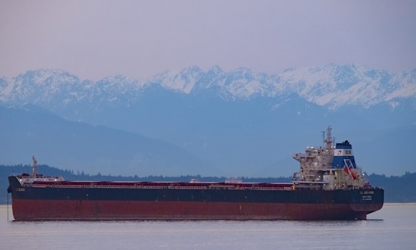 A large cargo ship is sailing across a calm body of water with a backdrop of snow-capped mountains and a hazy sky. The vessel is dark with a red hull and various structures on the deck, including a tall tower.