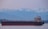 A large cargo ship is sailing across a calm body of water with a backdrop of snow-capped mountains and a hazy sky. The vessel is dark with a red hull and various structures on the deck, including a tall tower.