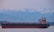 A large cargo ship is sailing across a calm body of water with a backdrop of snow-capped mountains and a hazy sky. The vessel is dark with a red hull and various structures on the deck, including a tall tower.