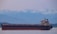 A large cargo ship is sailing across a calm body of water with a backdrop of snow-capped mountains and a hazy sky. The vessel is dark with a red hull and various structures on the deck, including a tall tower.