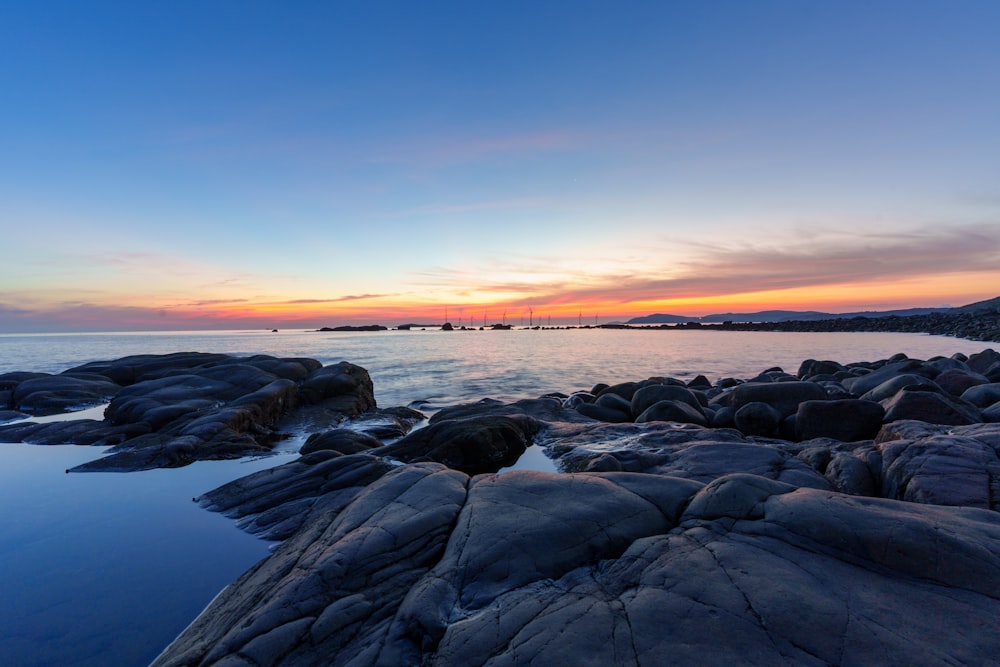 the sun is setting over the ocean with rocks in the foreground