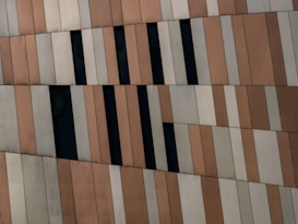 A close-up view of a building facade covered with vertical panels in shades of beige, brown, and grey. Some panels are replaced by black rectangular windows, creating a pattern. The panels appear to be slightly angled, adding to the visual interest.