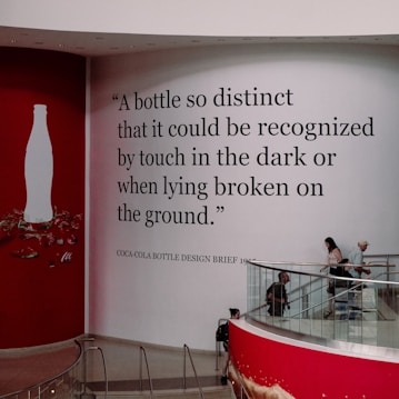 A large wall inside a modern building features an iconic silhouette of a bottle next to a prominent quote about the distinctiveness of the bottle's design. The text is in bold black lettering on a white background. Several people are walking down a curving staircase that is lined with red accents.