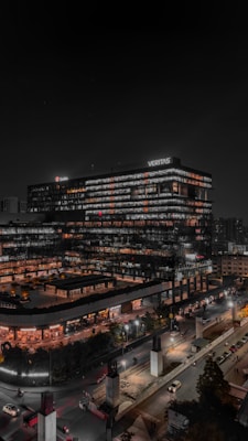 A modern multi-story office building with the name 'Veritas' prominently displayed on the top. The structure is illuminated with many lights, indicating activity or occupation during nighttime. The surrounding area includes a busy road with several cars and a construction site visible in the foreground.