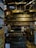 A well-stocked shop or bar area featuring a wide variety of bottles lined up on shelves that cover the back wall. The shelves are filled with different colors and types of bottles, suggesting a diverse selection of beverages. The foreground has more bottles and promotional materials that indicate some are on sale. Various signs and memorabilia, such as license plates and posters, decorate the space, adding a rustic or vintage appeal.