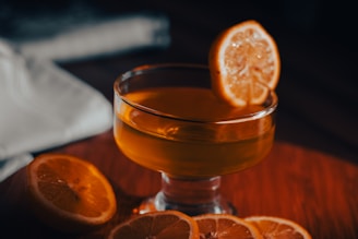 A glass of amber-colored liquid is garnished with a lemon slice and surrounded by additional lemon slices on a wooden surface. A white cloth is partially visible in the background.
