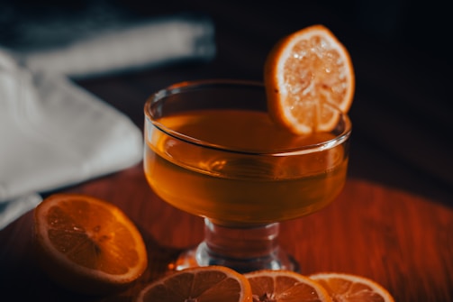 A glass of amber-colored liquid is garnished with a lemon slice and surrounded by additional lemon slices on a wooden surface. A white cloth is partially visible in the background.