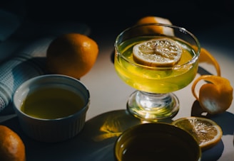 A glass dessert cup filled with lemon jelly, featuring a lemon slice on top. Nearby, whole lemons and a peeled lemon with its rind are arranged artistically. The setting is enhanced by a soft light that casts dramatic shadows around the objects.