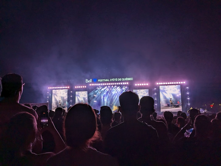A large crowd gathered in front of a brightly lit concert stage with multiple screens displaying live performances. The atmosphere is energetic with vibrant lights and music.