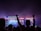 A large outdoor music festival at night features a stage with bright lights and a video screen displaying a musician. People in the crowd are silhouetted against the colorful lights, some with their arms raised in excitement.