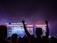 A large outdoor music festival at night features a stage with bright lights and a video screen displaying a musician. People in the crowd are silhouetted against the colorful lights, some with their arms raised in excitement.