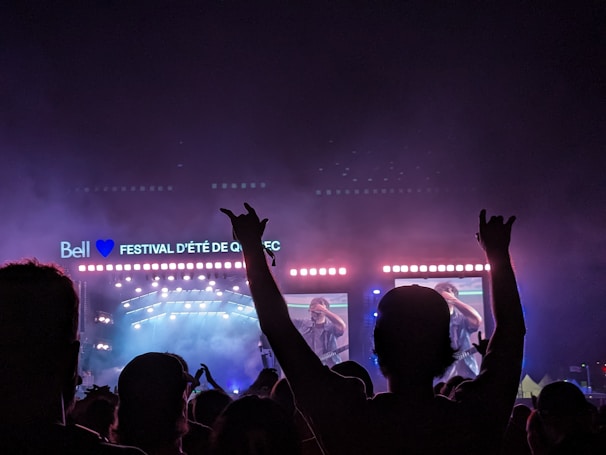 A large outdoor music festival at night features a stage with bright lights and a video screen displaying a musician. People in the crowd are silhouetted against the colorful lights, some with their arms raised in excitement.