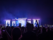 A large crowd is gathered in front of a brightly lit stage during a music festival. On the stage, there are large screens showing a musician playing a guitar. The night sky is visible above the stage, and various lighting effects create an atmospheric backdrop.
