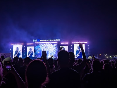 A large crowd is gathered in front of a brightly lit stage during a music festival. On the stage, there are large screens showing a musician playing a guitar. The night sky is visible above the stage, and various lighting effects create an atmospheric backdrop.
