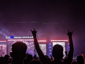 A large outdoor concert venue at night with a brightly lit stage featuring a live performance. The silhouette of the crowd is visible in the foreground, with some attendees raising their hands in excitement. Screens on either side of the stage display musicians playing.