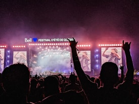 A nighttime outdoor concert scene with a large crowd in silhouette. The brightly lit stage displays several large screens showing a musician. Smoke effects and colorful lighting enhance the festive atmosphere. There's a sign above the stage reading 'Bell Festival D'été de Québec'.