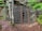 A secured entrance to a cave or mine is covered with a metal grid fence. The area is surrounded by rocks and vegetation, including a tree growing in front of the fence. The ground is earthy and covered in leaves and pine needles, suggesting a forested environment.