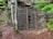 A secured entrance to a cave or mine is covered with a metal grid fence. The area is surrounded by rocks and vegetation, including a tree growing in front of the fence. The ground is earthy and covered in leaves and pine needles, suggesting a forested environment.