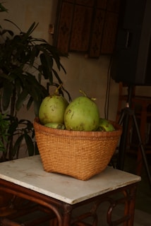 A wicker basket filled with several green coconuts is placed on a small wooden table with a marble top. In the background, there are lush green plants and decorative wooden panels on the wall.