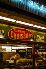 A food stall or counter with signage advertising 'Beef & Cheddar Croissant' and 'Italian Sausage'. The interior is dimly lit with hanging pendant lights. There's a food preparation area visible through the glass, containing kitchen equipment and a roll of paper towels.