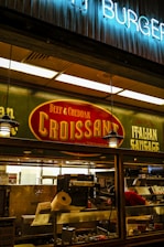 A food stall or counter with signage advertising 'Beef & Cheddar Croissant' and 'Italian Sausage'. The interior is dimly lit with hanging pendant lights. There's a food preparation area visible through the glass, containing kitchen equipment and a roll of paper towels.