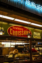 A food stall or counter with signage advertising 'Beef & Cheddar Croissant' and 'Italian Sausage'. The interior is dimly lit with hanging pendant lights. There's a food preparation area visible through the glass, containing kitchen equipment and a roll of paper towels.