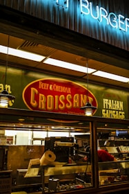 A food stall or counter with signage advertising 'Beef & Cheddar Croissant' and 'Italian Sausage'. The interior is dimly lit with hanging pendant lights. There's a food preparation area visible through the glass, containing kitchen equipment and a roll of paper towels.