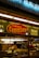 A food stall or counter with signage advertising 'Beef & Cheddar Croissant' and 'Italian Sausage'. The interior is dimly lit with hanging pendant lights. There's a food preparation area visible through the glass, containing kitchen equipment and a roll of paper towels.