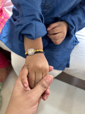 A child's hand adorned with a bracelet is being gently held by an adult hand. The bracelet is gold and silver with the text 'BRO' on it. The child is wearing a blue garment, and there is a hint of another person in pink clothing in the background.
