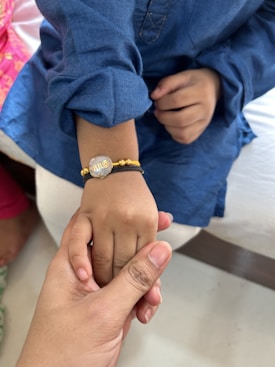 A child's hand adorned with a bracelet is being gently held by an adult hand. The bracelet is gold and silver with the text 'BRO' on it. The child is wearing a blue garment, and there is a hint of another person in pink clothing in the background.