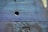 The image depicts a close-up view of a textured cement wall with an uneven surface. The wall features various shades of blue and subtle hues of purple and green, with small cracks and a noticeable indented area in the center.