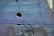 The image depicts a close-up view of a textured cement wall with an uneven surface. The wall features various shades of blue and subtle hues of purple and green, with small cracks and a noticeable indented area in the center.
