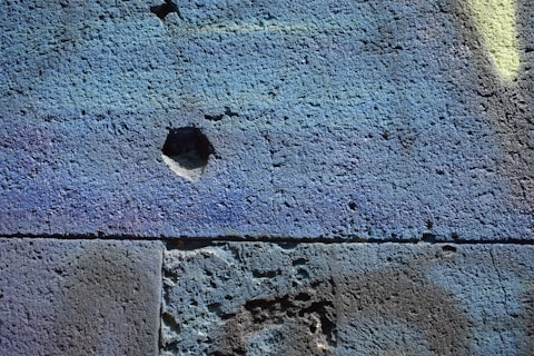 The image depicts a close-up view of a textured cement wall with an uneven surface. The wall features various shades of blue and subtle hues of purple and green, with small cracks and a noticeable indented area in the center.