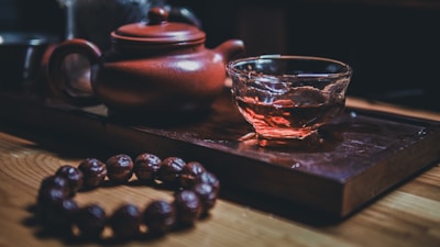 A traditional setup featuring a red clay teapot and a clear glass teacup filled with tea on a wooden tray. In the foreground, there is a bead bracelet arranged on a smooth wooden surface, with warm ambient lighting casting soft shadows.
