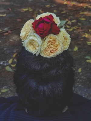 A top bun hairstyle adorned with a bouquet of flowers, featuring a vivid red rose surrounded by creamy white roses. The background is a textured surface with scattered yellow leaves, suggesting an outdoor setting.