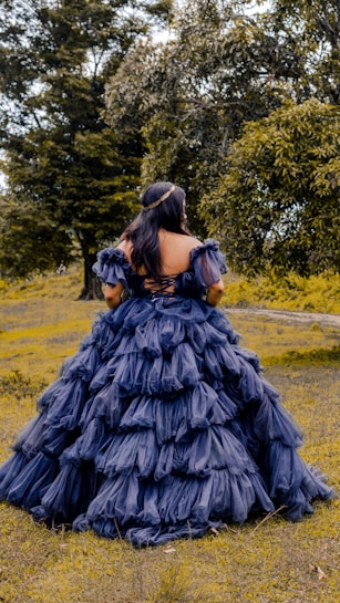 A person stands in a natural setting, wearing a voluminous, layered gown in a deep blue color. The dress features ruffled details and an off-the-shoulder design, with the person facing away, showcasing their long hair adorned with a simple headband. Surrounding the individual are lush green trees and grass, creating a serene and picturesque atmosphere.