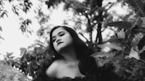 A black and white portrait of a person with long dark hair, posed amidst leafy foliage. The background is filled with blurred trees, creating a natural outdoor setting.