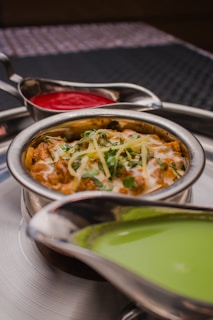 A stainless steel bowl filled with a creamy curry is garnished with grated cheese and cilantro, surrounded by two separate sauce holders containing vibrant red and green sauces. The presentation is on a metallic tray, creating an appetizing and colorful display.