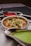 A stainless steel bowl filled with a creamy curry is garnished with grated cheese and cilantro, surrounded by two separate sauce holders containing vibrant red and green sauces. The presentation is on a metallic tray, creating an appetizing and colorful display.
