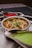 A stainless steel bowl filled with a creamy curry is garnished with grated cheese and cilantro, surrounded by two separate sauce holders containing vibrant red and green sauces. The presentation is on a metallic tray, creating an appetizing and colorful display.