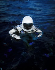 An astronaut in a spacesuit is partially submerged in dark water. The suit is white with blue and black details, featuring a NASA patch, and the helmet visor reflects light. The surrounding water is deep blue with subtle color variations.