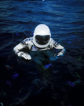 An astronaut in a spacesuit is partially submerged in dark water. The suit is white with blue and black details, featuring a NASA patch, and the helmet visor reflects light. The surrounding water is deep blue with subtle color variations.